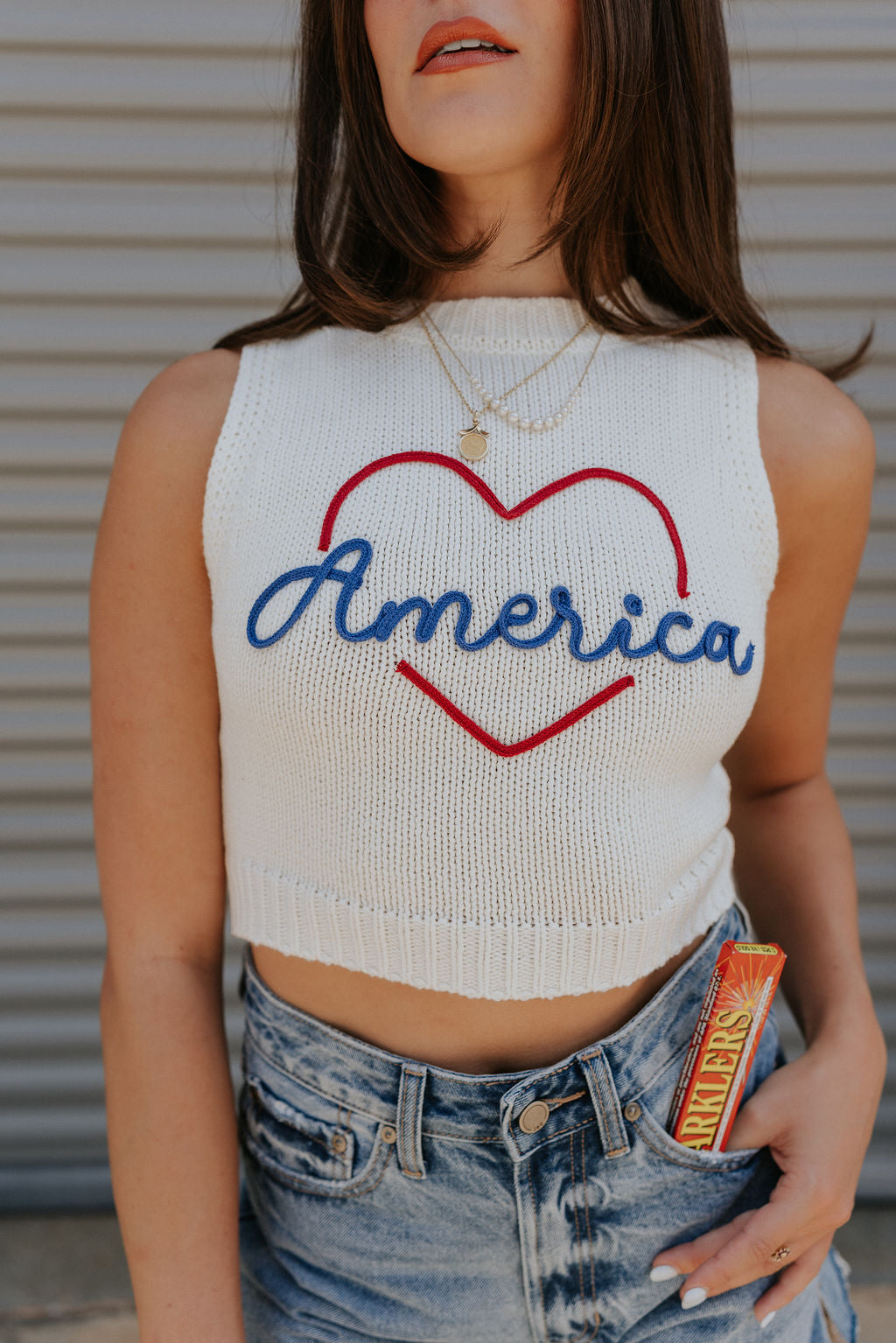 close up view of female model wearing the Love America Cream Knit Tank which features Cream Knit Fabric, Round Neckline, Sleeveless and Red Stitch Heart, Says America in blue stitch writing