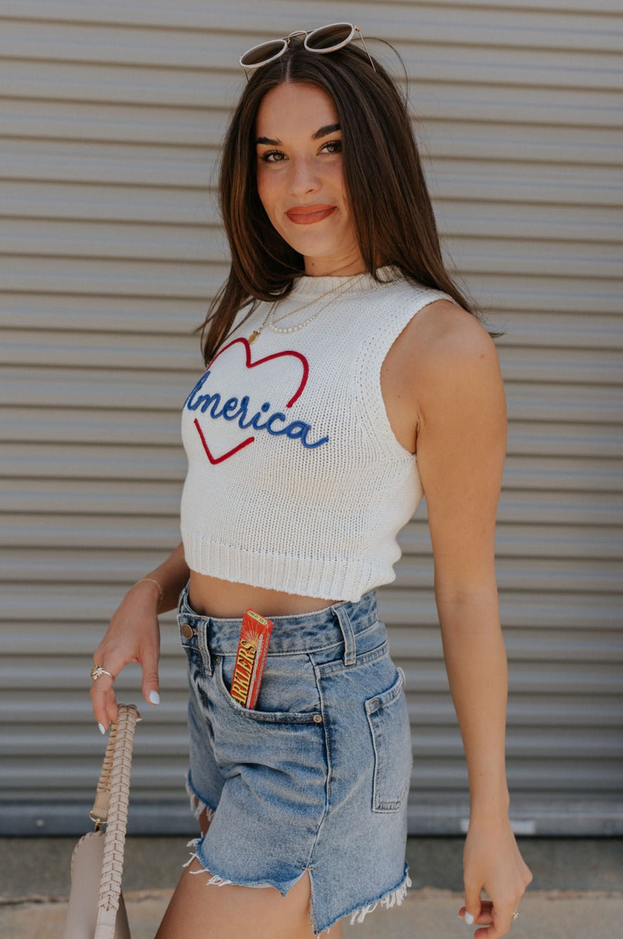 side view of female model wearing the Love America Cream Knit Tank which features Cream Knit Fabric, Round Neckline, Sleeveless and Red Stitch Heart, Says America in blue stitch writing