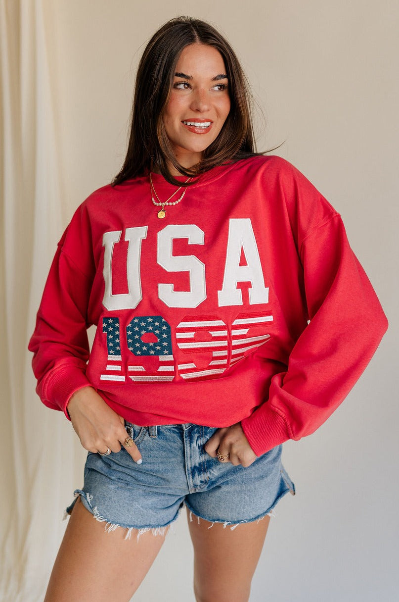 front view of female model wearing the USA 1996 Red Long Sleeve Sweatshirt which features Lightweight Red Fabric, Round Neckline, Long Sleeves, Ribbed Hem and USA 1996 graphic with white, red and blue stitch design