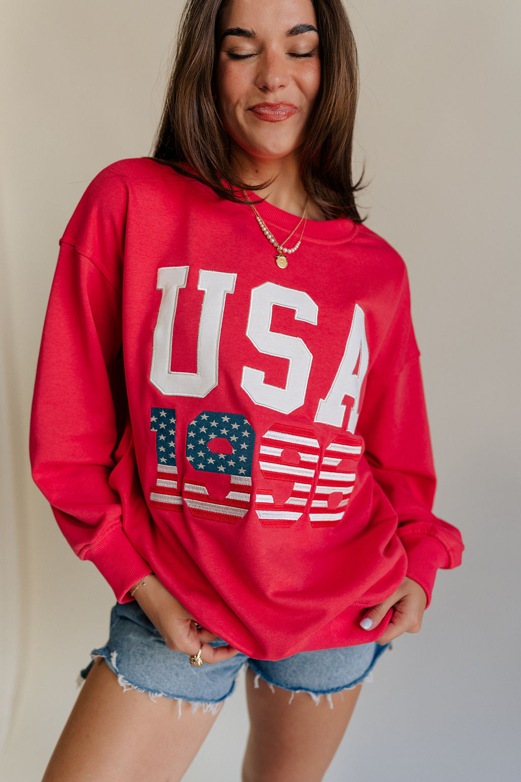 close up view of female model wearing the USA 1996 Red Long Sleeve Sweatshirt which features Lightweight Red Fabric, Round Neckline, Long Sleeves, Ribbed Hem and USA 1996 graphic with white, red and blue stitch design