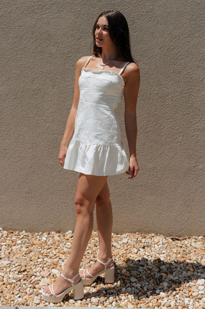 full body view of female model wearing the Faye White Floral Ruffle Mini Dress which features White Lightweight Fabric, Monochrome Floral & Paisley Design, Ruffle Hem Skirt, White Shorts Lining, Sweetheart Ruffle Neckline, Adjustable Straps and Monochrome Back Zipper with Hook Closure