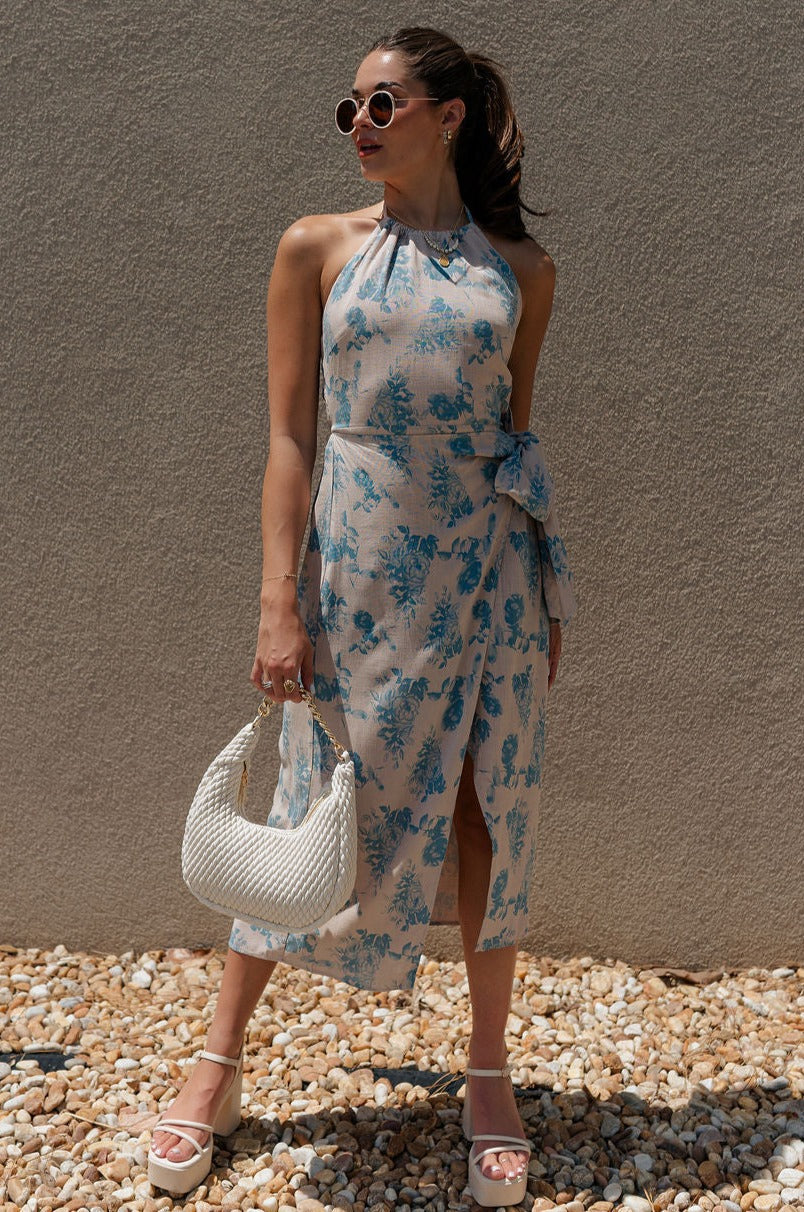 full body view of female model wearing the Mara Sand & Vintage Blue Floral Midi Dress which features Sand Linen Fabric, Slate Blue Floral Design, Midi Length, Slit Design, Waist Tie Detail, Halter Neckline with Tie Closure and Smocked Back