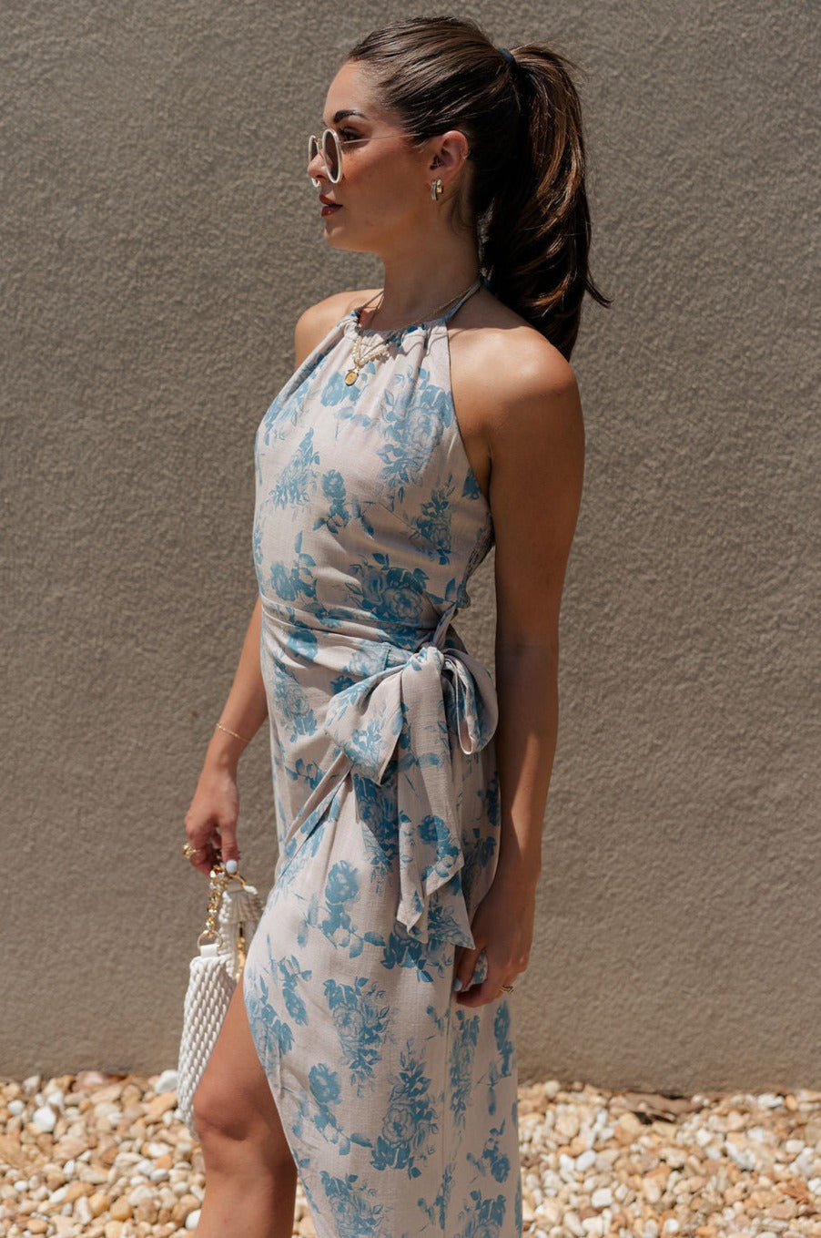 side view of female model wearing the Mara Sand & Vintage Blue Floral Midi Dress which features Sand Linen Fabric, Slate Blue Floral Design, Midi Length, Slit Design, Waist Tie Detail, Halter Neckline with Tie Closure and Smocked Back