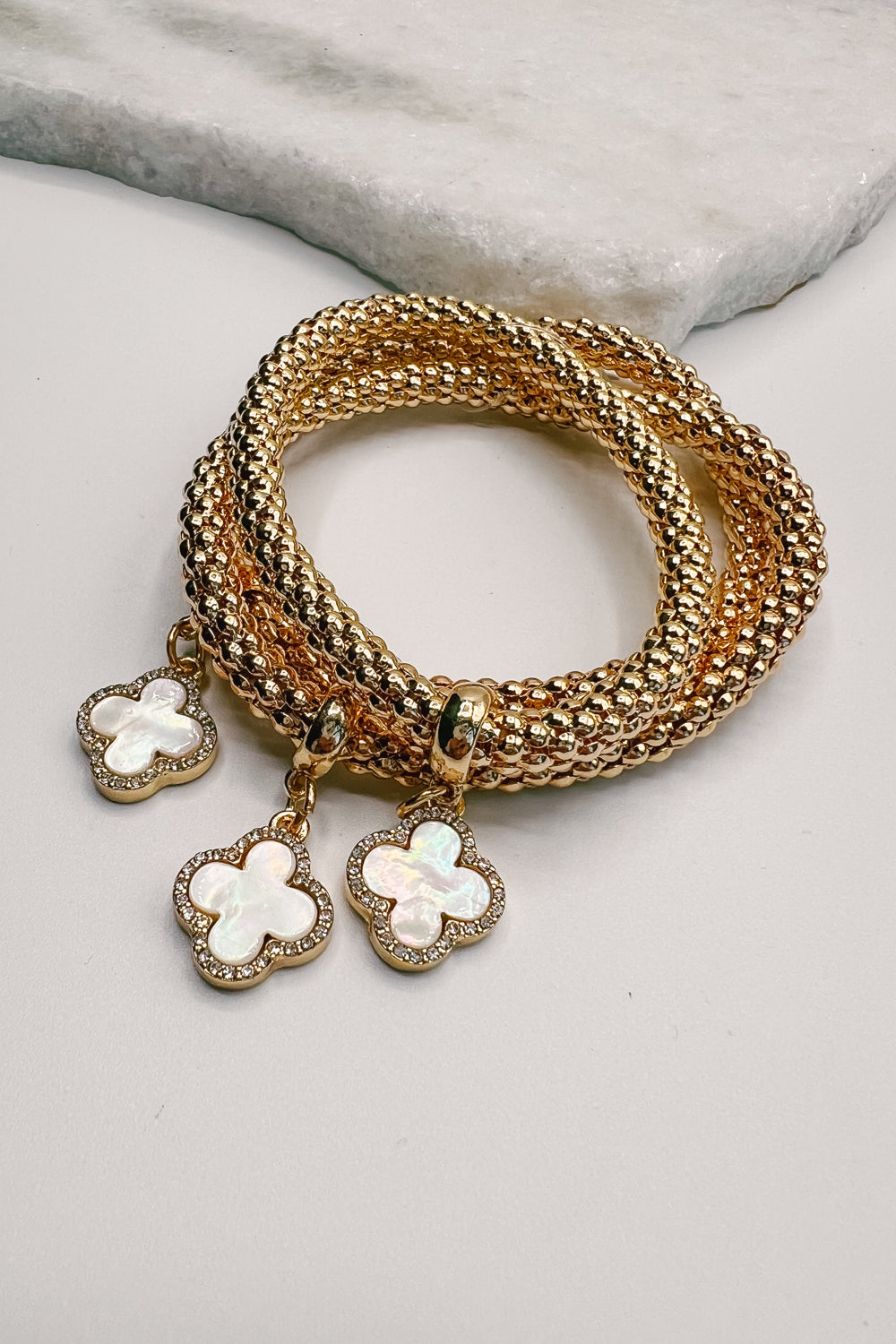 A close-up of the Clove Bracelet Set is shown. The set is made of three gold cobra bracelets with clover charms. 