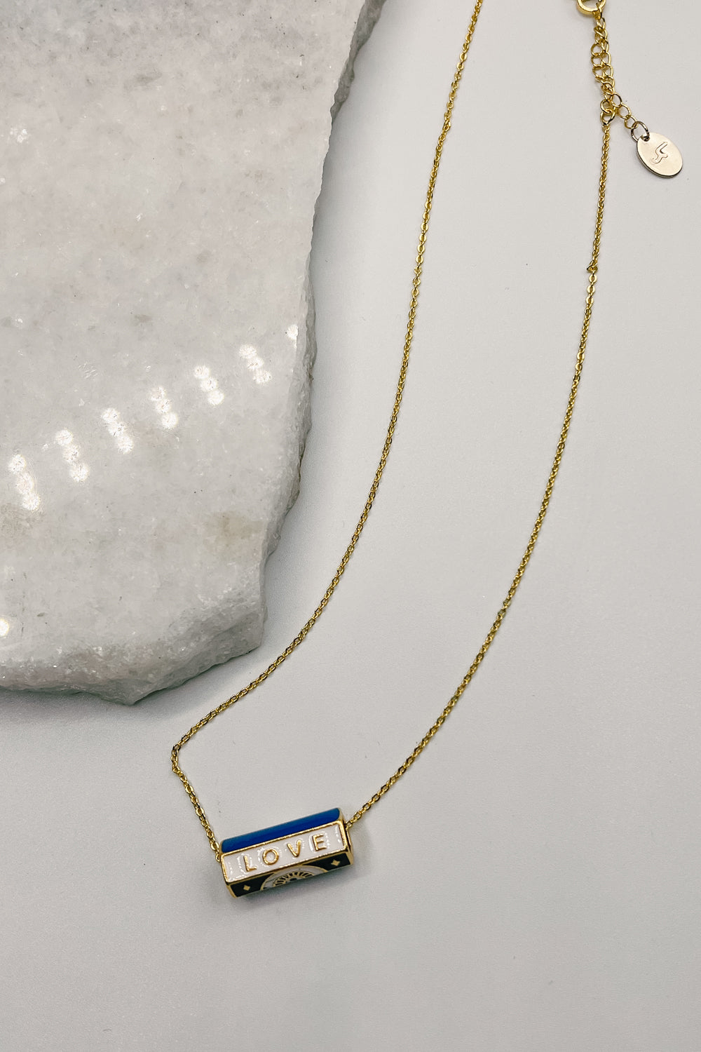 Image shows Love White & Blue Cylinder Necklace against a white background. Necklace has 6 sided cylinder - 2 white sides that say love, 2 blue sides, and 2 black sides with a geometric design.