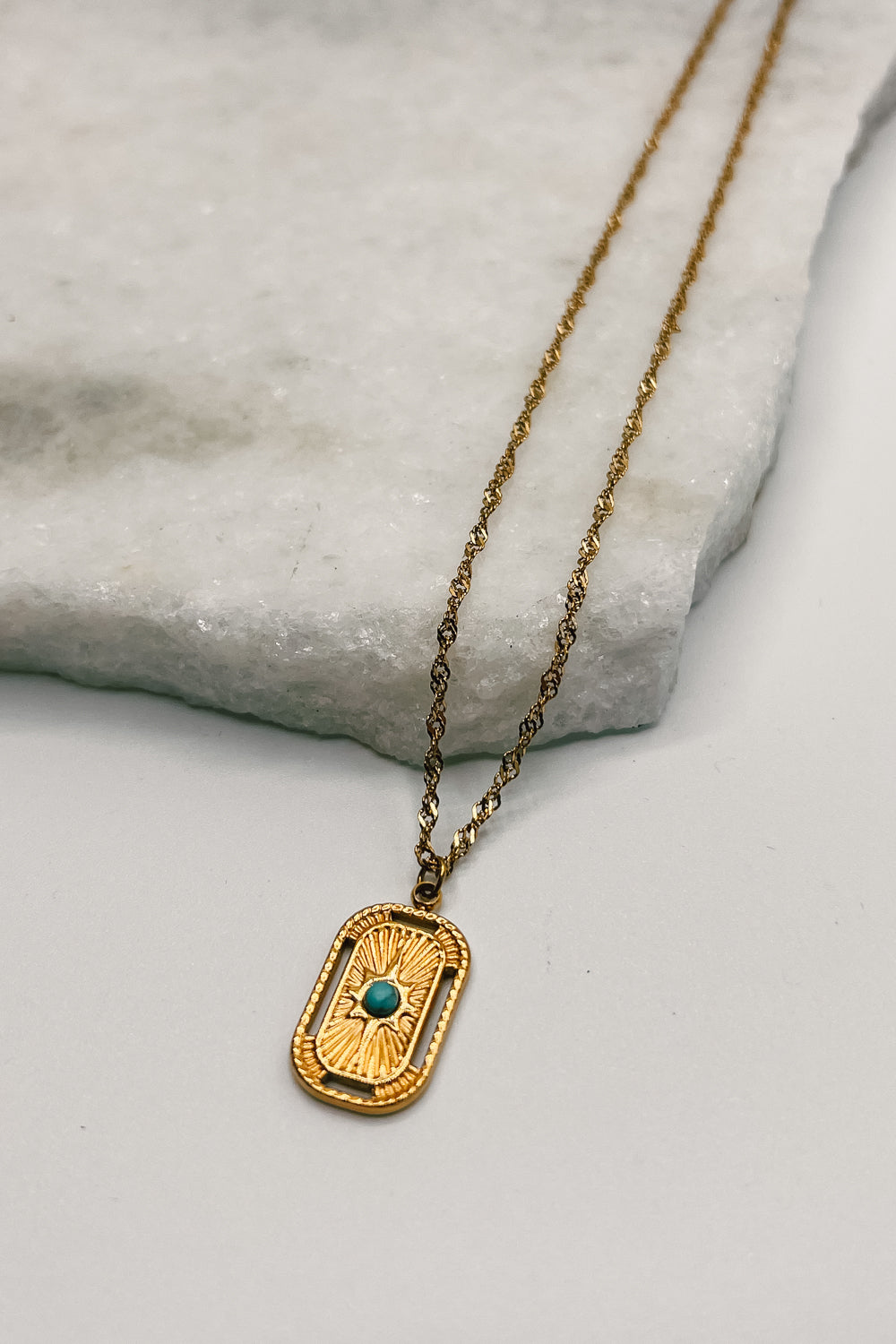 Image shows Liora Gold & Turquoise Medallion Necklace against a white marble background. Necklace has gold chain and a gold rectangle medallion with a turquoise stone.