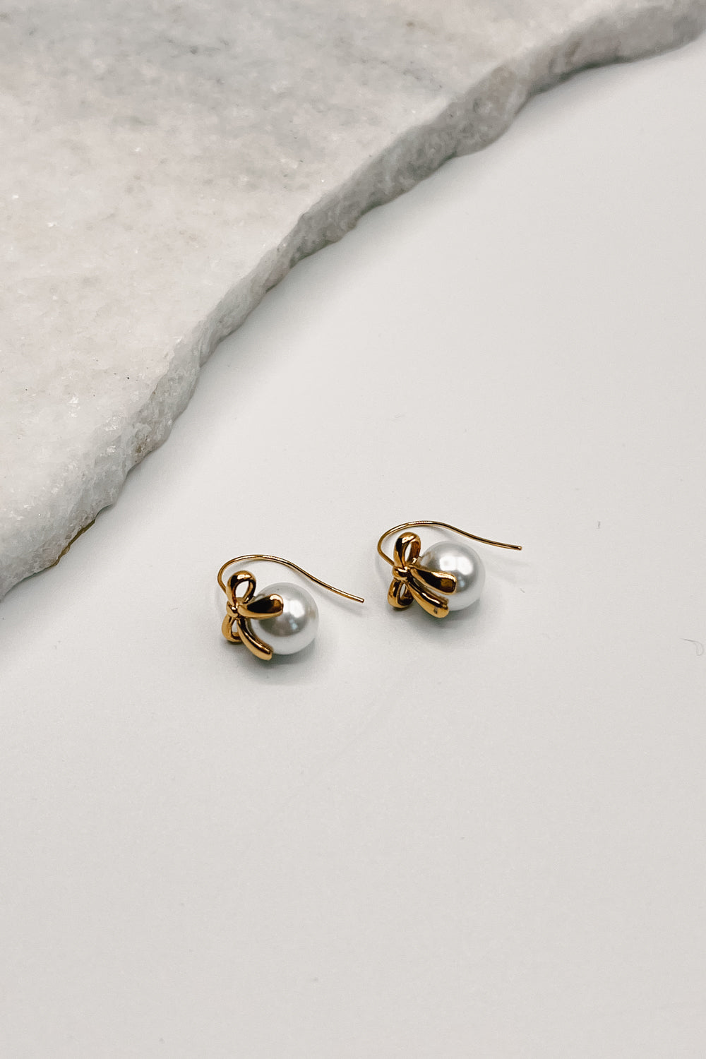 flat lay view of the Lara Pearl & Gold Bow Earring which features small gold dangle earrings with pearl and bow detail