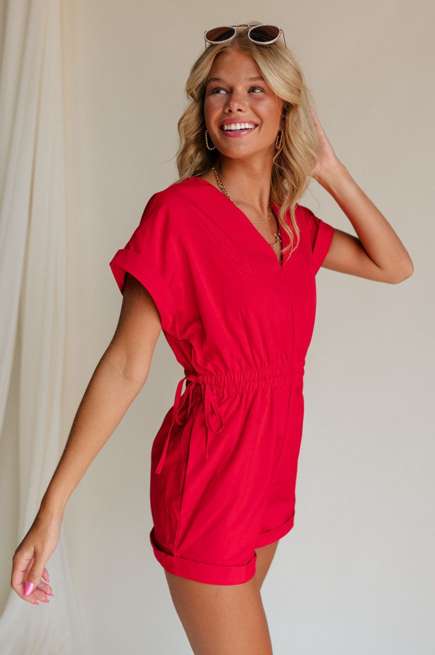 side view of female model wearing the Margot Tie Waist Short Sleeve Romper which features Elastic Waistband with Ties, Lightweight Fabric, V Neckline, Short Sleeves and Side Pockets. the romper is available in red and blue.