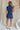 back view of female model wearing the Lila Navy Denim Button-Up Romper which features Navy Blue Washed Fabric, Two Front Pockets, Two Back Pockets, Front Zipper with Button Up Closure, Collared Neckline and Sleeveless