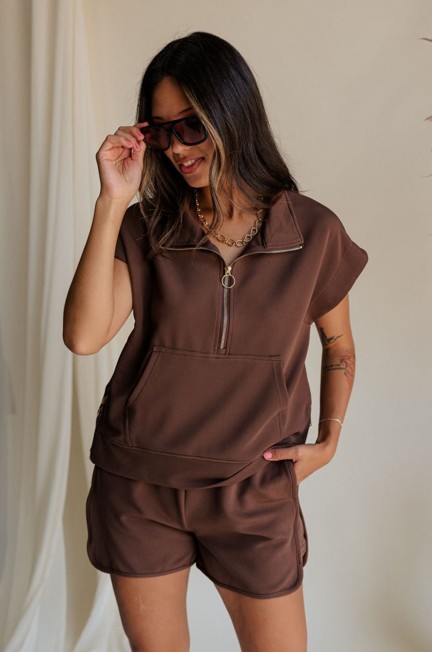 front view of female model wearing the Thea Brown Ribbed Sleeveless Top which features Brown Ribbed Fabric, One Large Front Pocket, Quarter Zip Up, High Neckline, Gold Zipper Side Details and Sleeveless