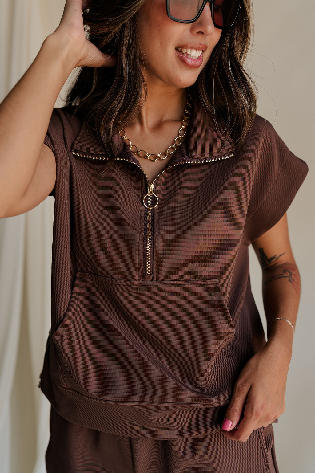close up view of female model wearing the Thea Brown Ribbed Sleeveless Top which features Brown Ribbed Fabric, One Large Front Pocket, Quarter Zip Up, High Neckline, Gold Zipper Side Details and Sleeveless