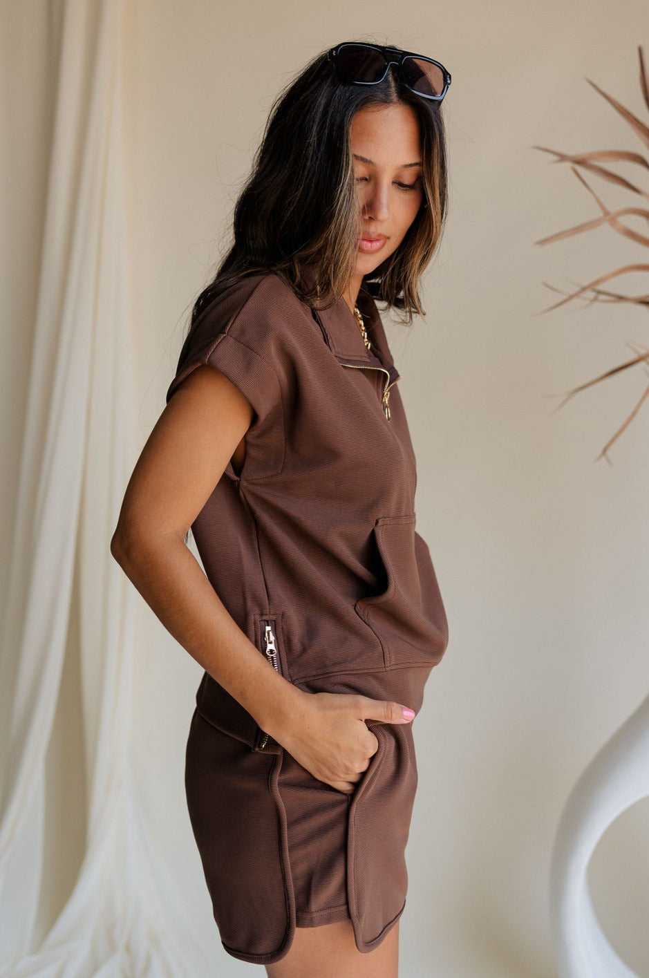 side view of female model wearing the Thea Brown Ribbed Sleeveless Top which features Brown Ribbed Fabric, One Large Front Pocket, Quarter Zip Up, High Neckline, Gold Zipper Side Details and Sleeveless