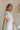 Upper body side view of female model wearing the Elysia Short Sleeve Ruffle Top that has cream fabric, ruffle details, front pleating, and slit sleeves with ties.