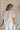 Back view of female model wearing the Elysia Short Sleeve Ruffle Top that has cream fabric, ruffle details, front pleating, and slit sleeves with ties.