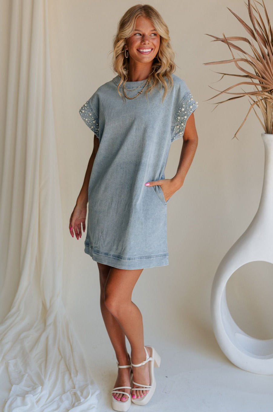 full body view of female model wearing the Raelyn Light Denim Pearl & Rhinestone Mini Dress which features Light Denim Wash Fabric, Side Pockets, Pearl and Rhinestone Details, Round Neckline, Short Sleeves and Back Zipper with Hook Closure