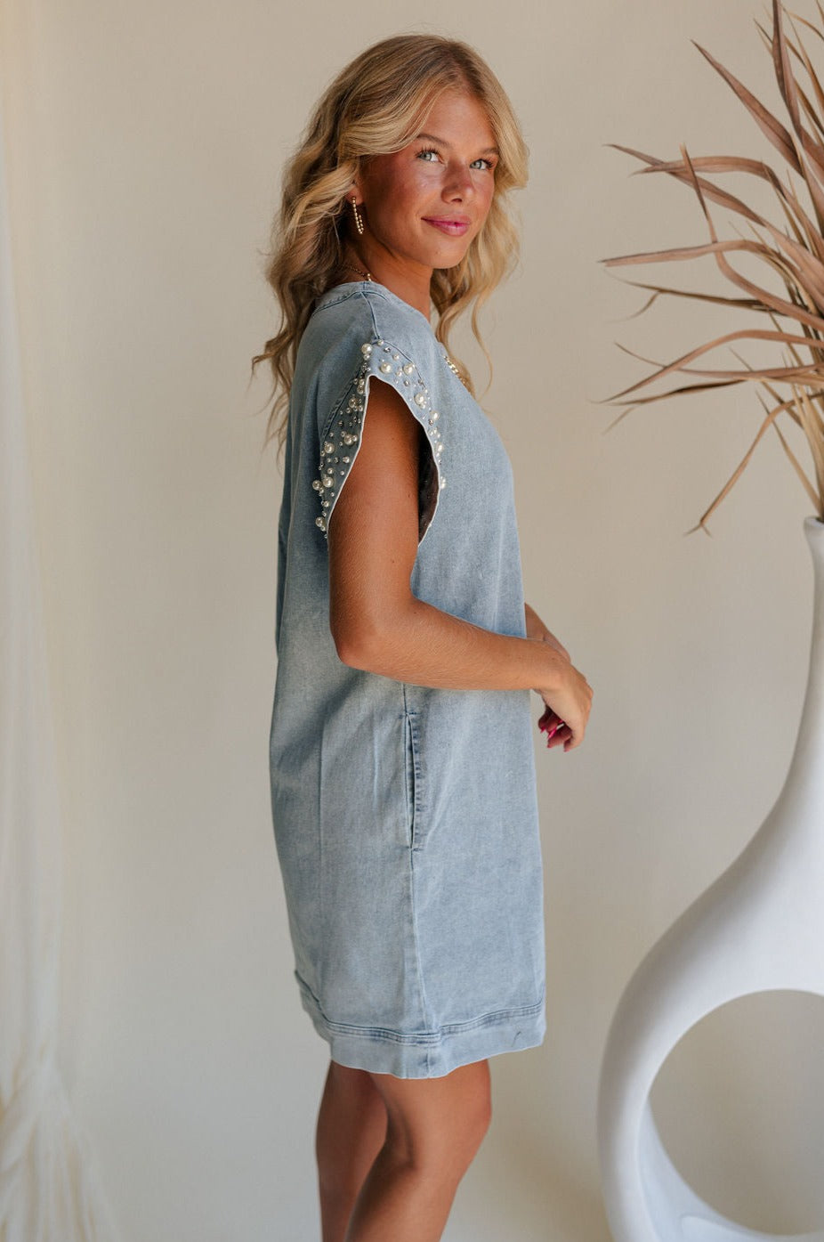 side view of female model wearing the Raelyn Light Denim Pearl & Rhinestone Mini Dress which features Light Denim Wash Fabric, Side Pockets, Pearl and Rhinestone Details, Round Neckline, Short Sleeves and Back Zipper with Hook Closure