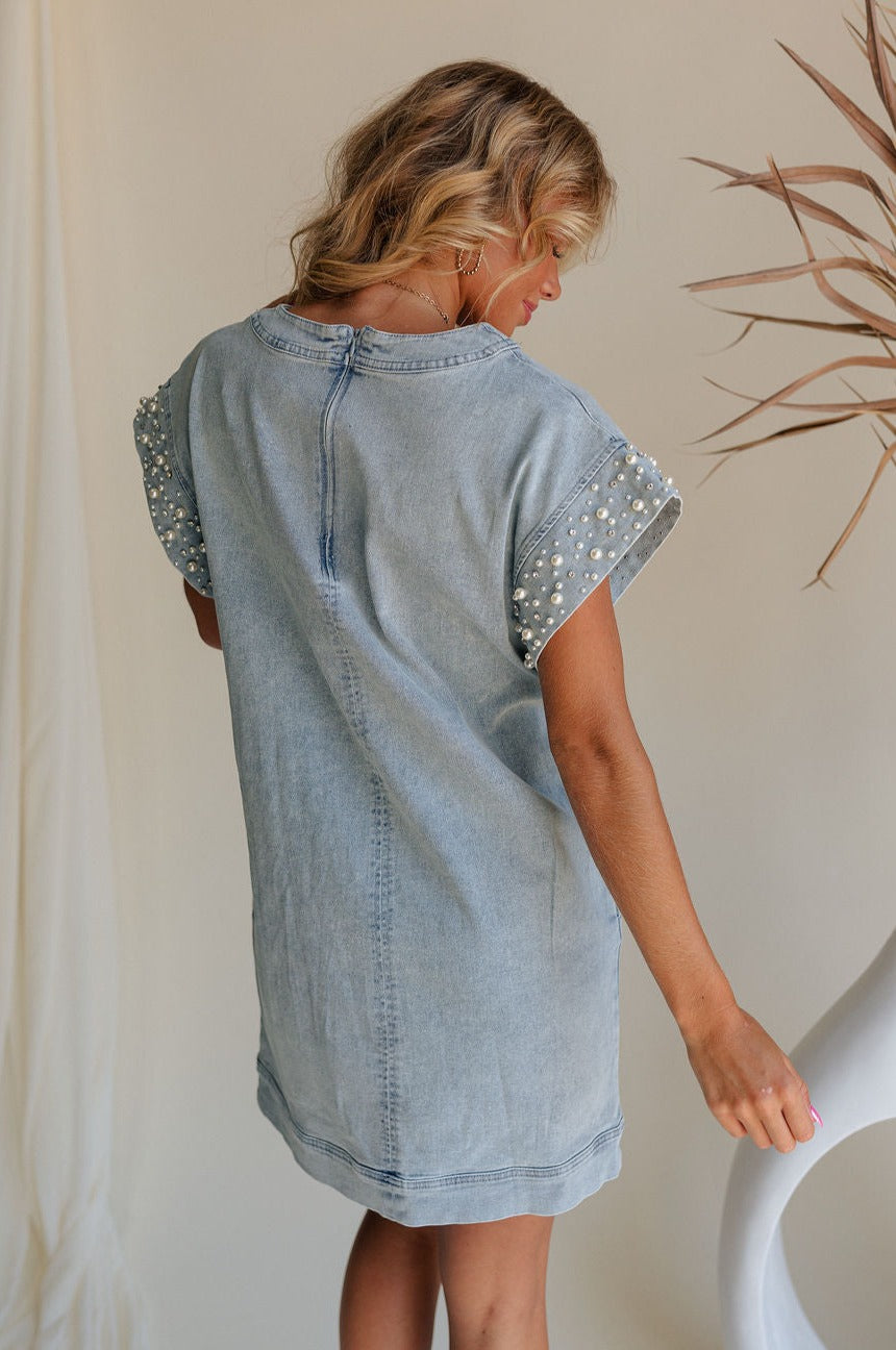 back view of female model wearing the Raelyn Light Denim Pearl & Rhinestone Mini Dress which features Light Denim Wash Fabric, Side Pockets, Pearl and Rhinestone Details, Round Neckline, Short Sleeves and Back Zipper with Hook Closure