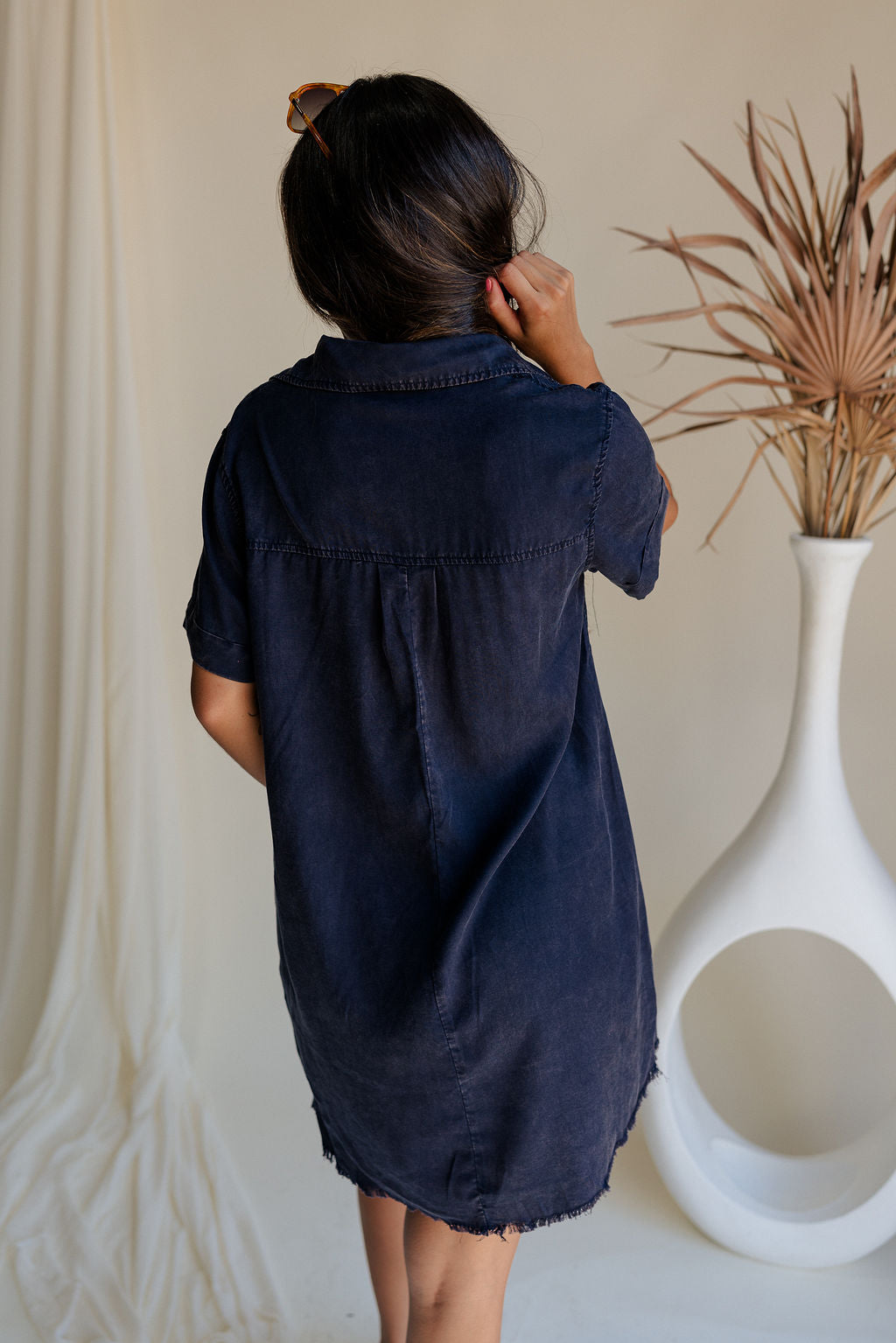 back view of female model wearing the Leighton Navy Fray Short Sleeve Mini Dress which featuresNavy Blue Tencel Fabric, Fray Hem Details, Mini Length, Quarter Button-Up Closure, Collared Neckline, Short Sleeves and Left Front Chest Pocket