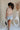 Full body back view of female model wearing the Elysia Short Sleeve Ruffle Top in Taupe that has taupe fabric, ruffle details, front pleating, and slit sleeves with ties.