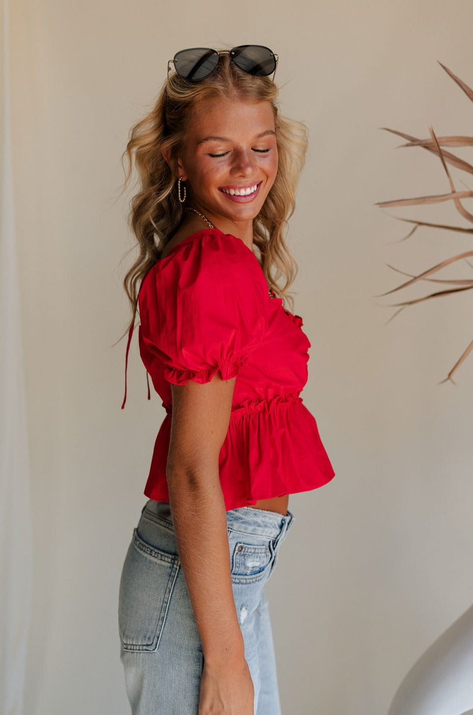 Upper body side view of female model wearing the Monet Red Puff Sleeve Crop Top that has red fabric, puff short sleeves, a cropped flared waist, and an open back with a tie.