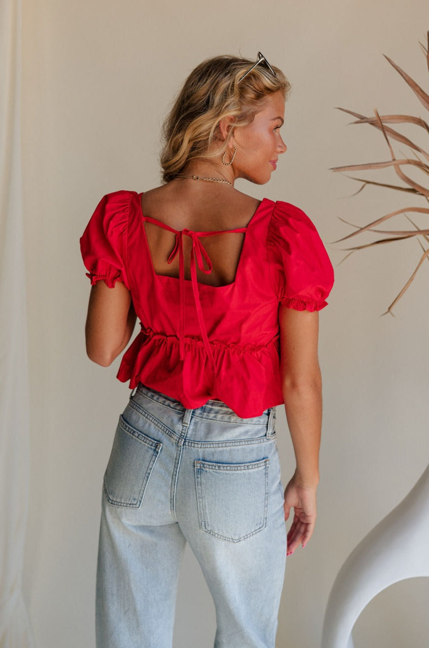 Upper body back view of female model wearing the Monet Red Puff Sleeve Crop Top that has red fabric, puff short sleeves, a cropped flared waist, and an open back with a tie.