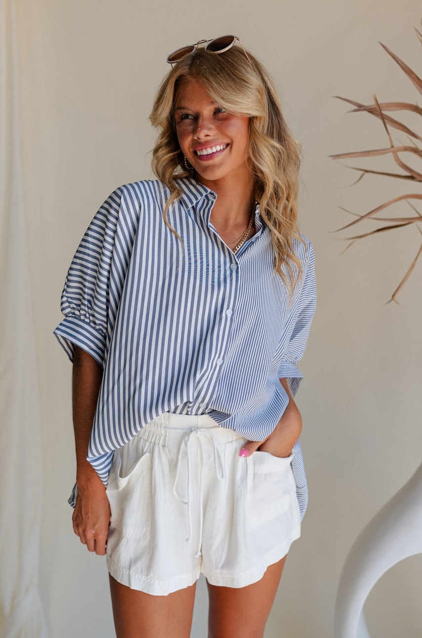 Front view of female model wearing the Liora Striped Short Sleeve Button Up Top in blue that has blue and white stripes, a button up front, a collar, and short sleeves. Front tucked into white shorts.