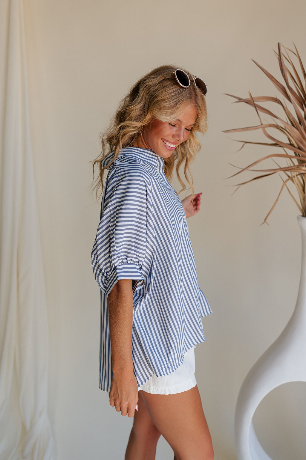 Side view of female model wearing the Liora Striped Short Sleeve Button Up Top in blue that has blue and white stripes, a button up front, a collar, and short sleeves.