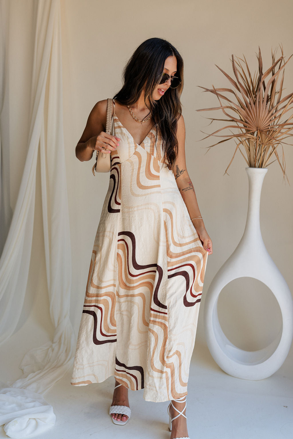 Full body front view of female model wearing the Wren Neutral Swirl Print Maxi Dress that has cream fabric with a taupe and brown swirl print, a v neck, and an open back with ties.