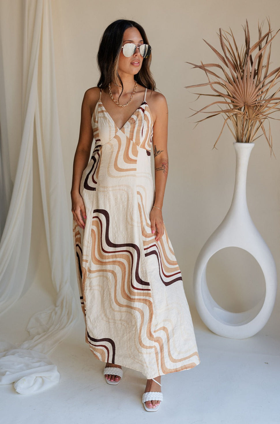Full body front view of female model wearing the Wren Neutral Swirl Print Maxi Dress that has cream fabric with a taupe and brown swirl print, a v neck, and an open back with ties.