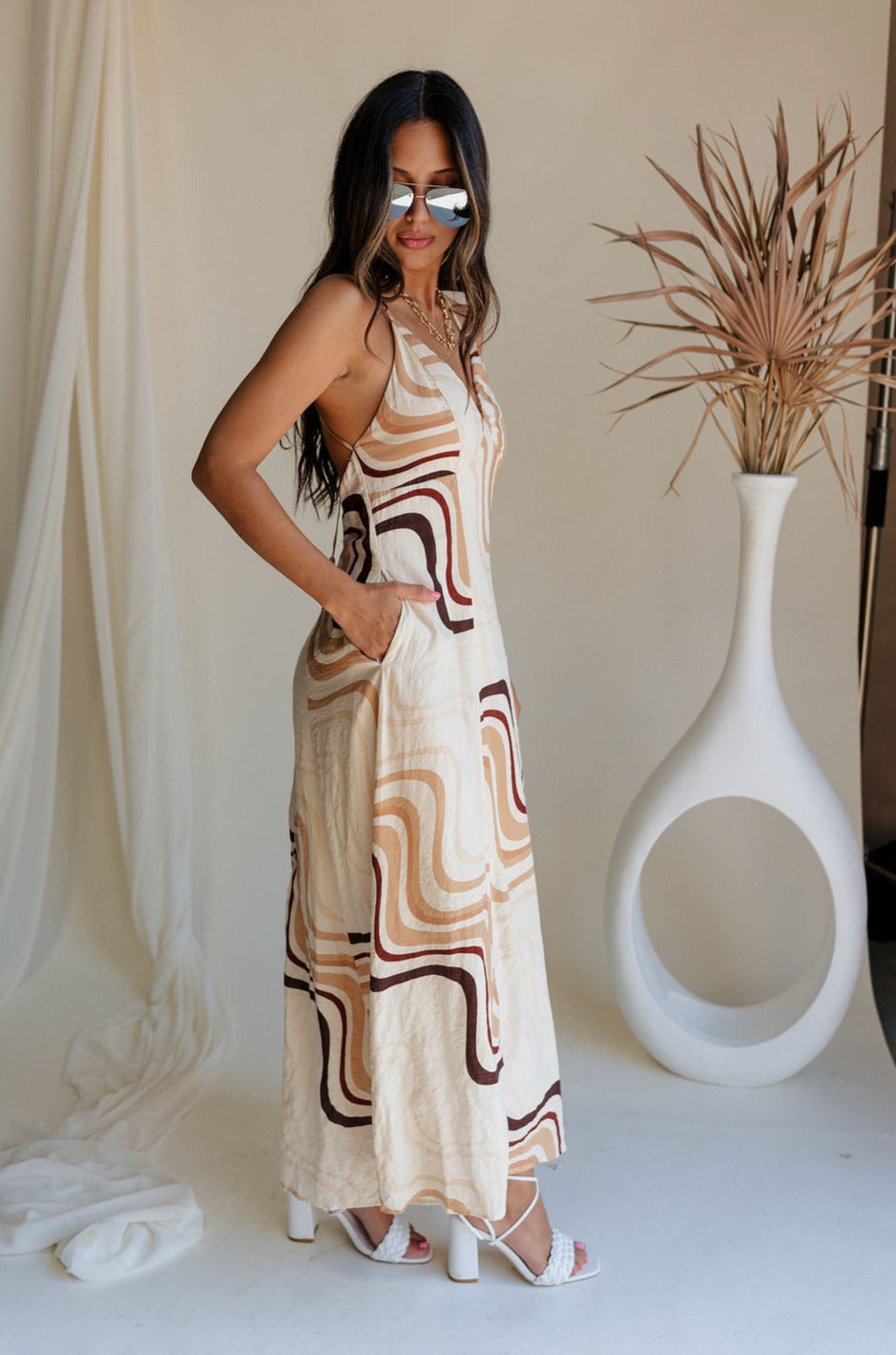 Full body side view of female model wearing the Wren Neutral Swirl Print Maxi Dress that has cream fabric with a taupe and brown swirl print, a v neck, and an open back with ties.