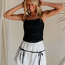 Upper body front view of female model wearing the Nina Bow Detail Mini Skirt in white. Skirt has white fabric with black bows in middle. Paired with black tank top.