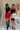 Full body front view of two female models wearing the Nina Bow Detail Mini Skirt in red and white, that has black bows in the middle, and an elastic waist. Worn with black top and black boots and black heels..