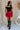 Full body front view of female model wearing the Nina Bow Detail Mini Skirt in Red that has red fabric, black bows in the middle, and an elastic waist. Worn with black top and black boots.