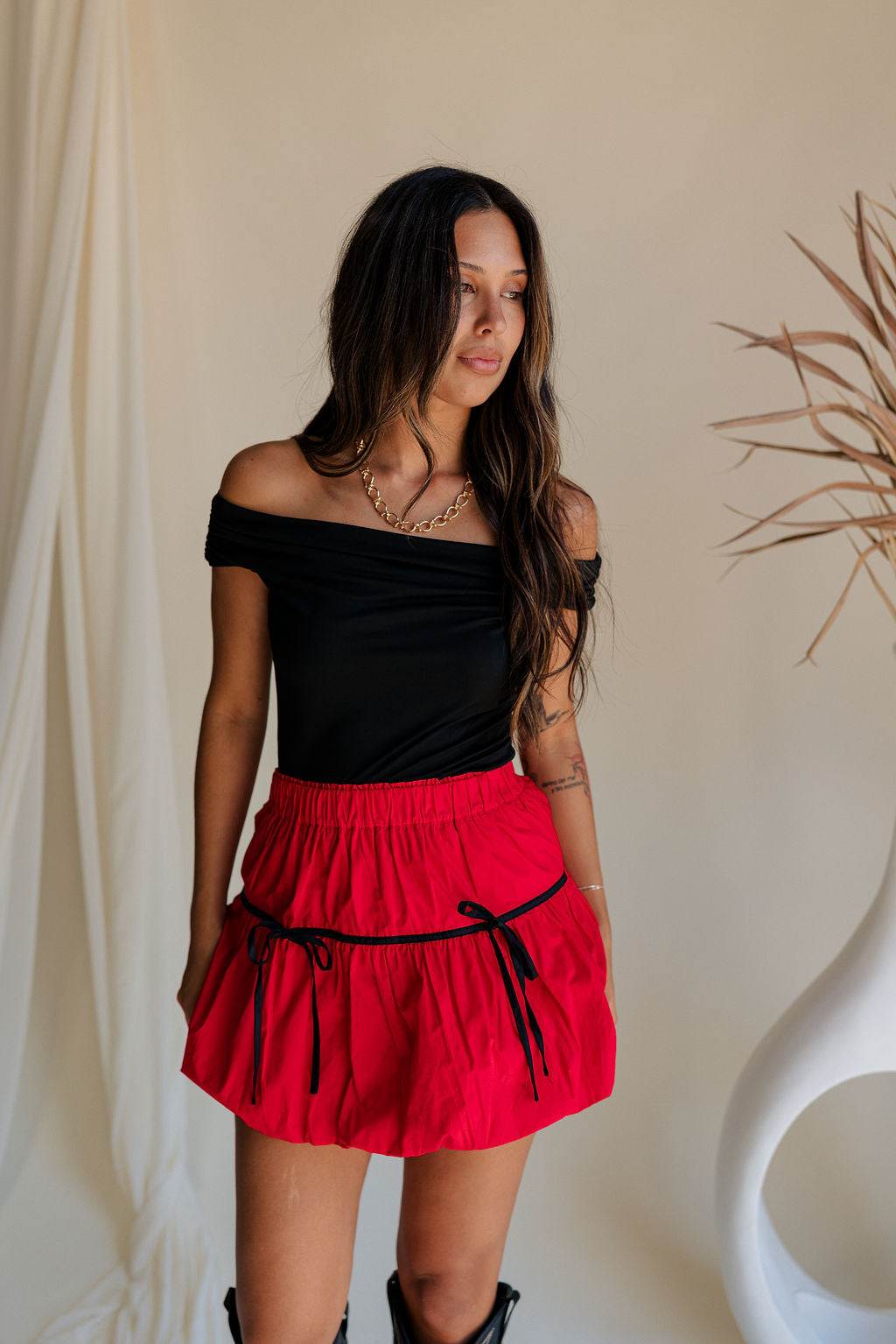 Upper body front view of female model wearing the Nina Bow Detail Mini Skirt in Red that has red fabric, black bows in the middle, and an elastic waist. Worn with black top and black boots.