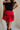 Close-up front view of female model wearing the Nina Bow Detail Mini Skirt in Red that has red fabric, black bows in the middle, and an elastic waist. Worn with black top and black boots.