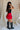 Full body side view of female model wearing the Nina Bow Detail Mini Skirt in Red that has red fabric, black bows in the middle, and an elastic waist. Worn with black top and black boots.