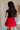 Upper body back view of female model wearing the Nina Bow Detail Mini Skirt in Red that has red fabric, black bows in the middle, and an elastic waist. Worn with black top and black boots.