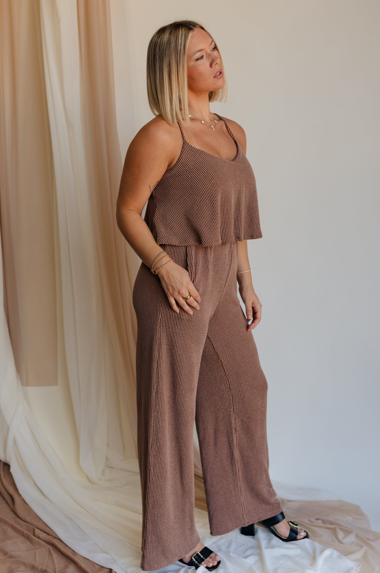 Full body side view of model wearing Kaylee Brown Ribbed Jumpsuit. This is a brown ribbed jumpsuit, with ruffle upper, and adjustable straps.