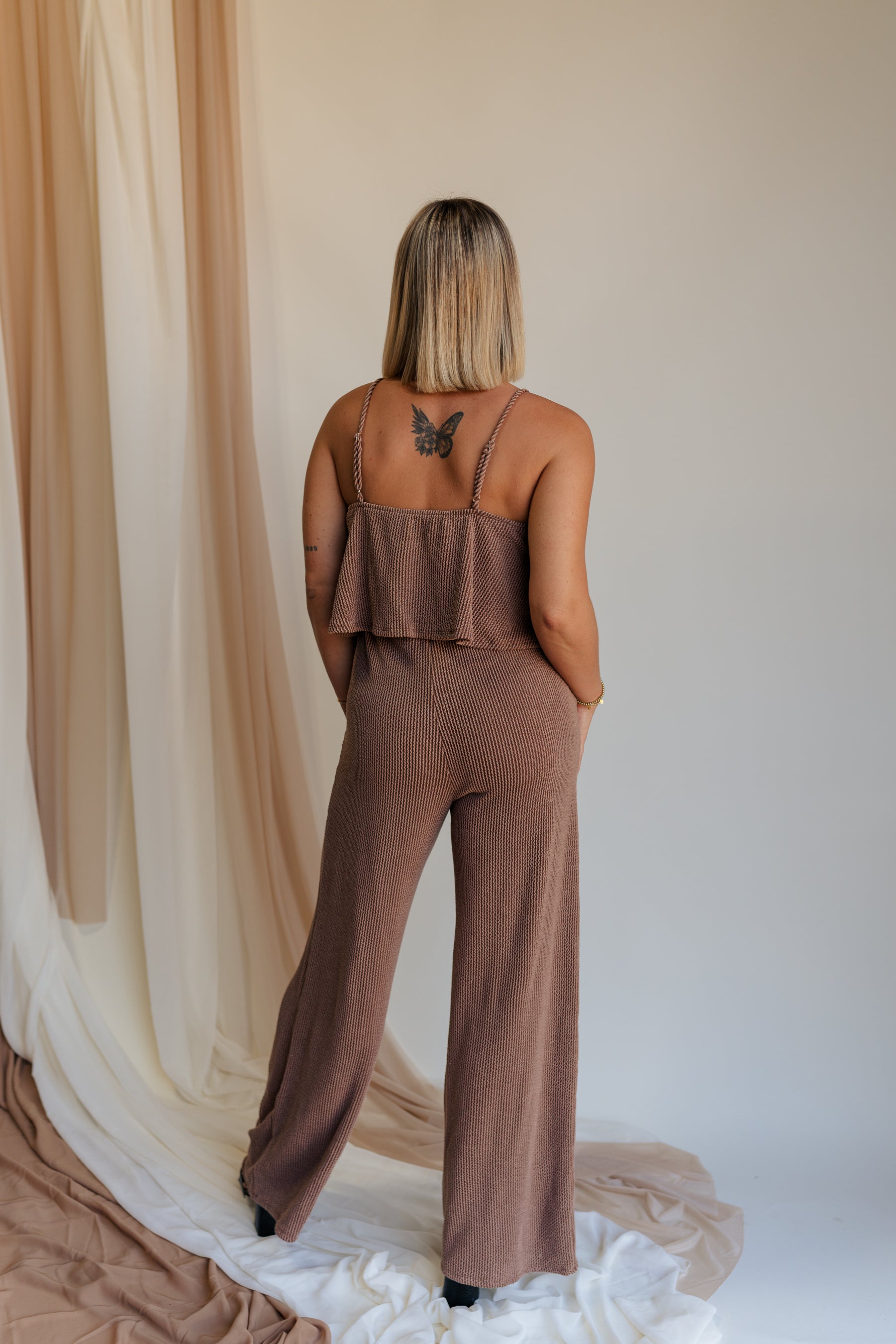 Full body back view of model wearing Kaylee Brown Ribbed Jumpsuit. This is a brown ribbed jumpsuit, with ruffle upper, and adjustable straps.