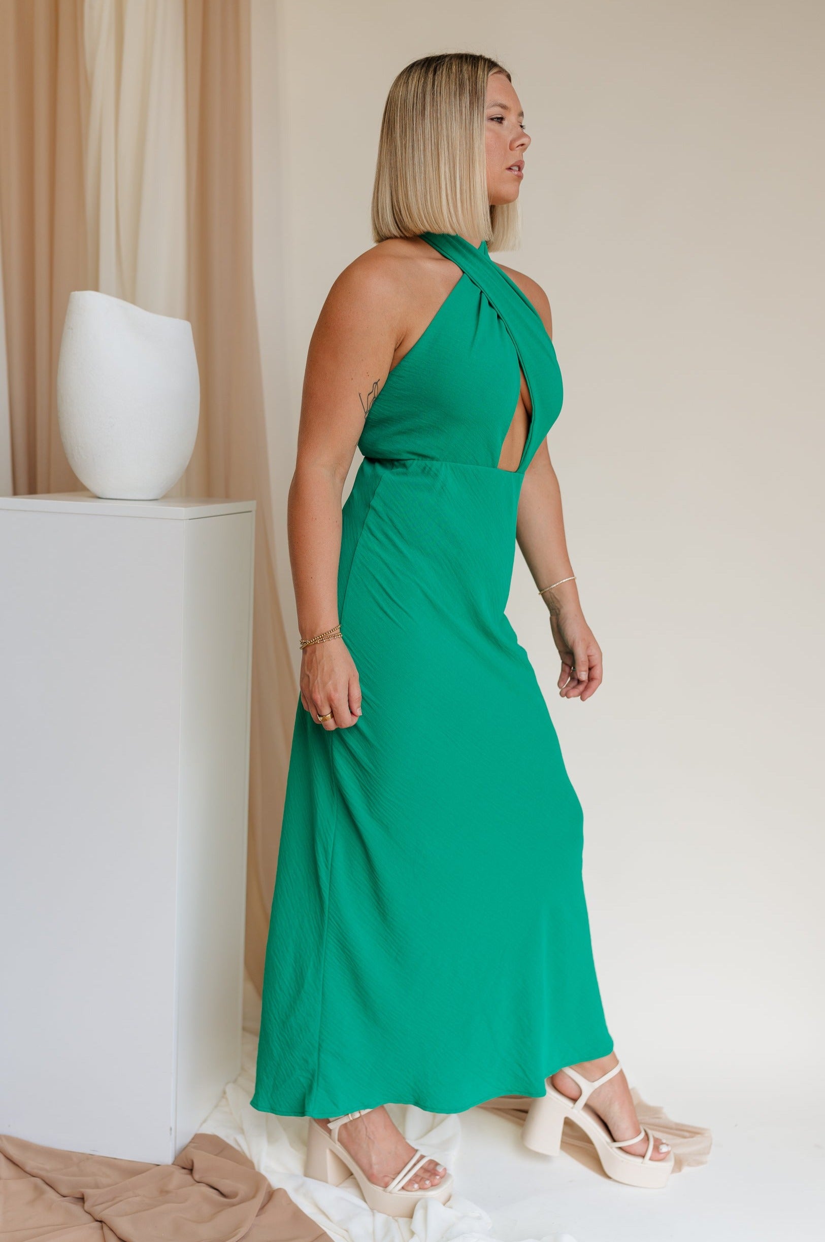 Side view of model wearing Madelyn Green Halter Maxi Dress. This is a green maxi dress with a halter criss cross neckline.