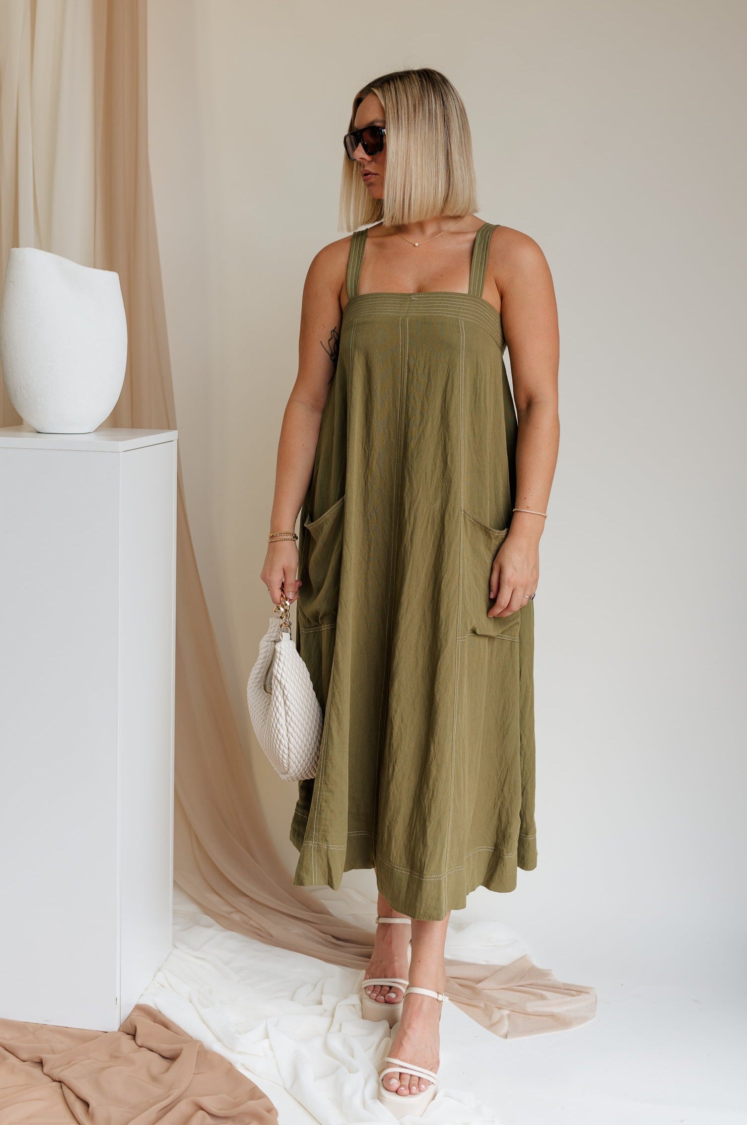Full body view of model wearing Melanie Olive Midi Dress. This is an olive midi dress, with white stitch details, midi length, pockets, and adjustable straps.