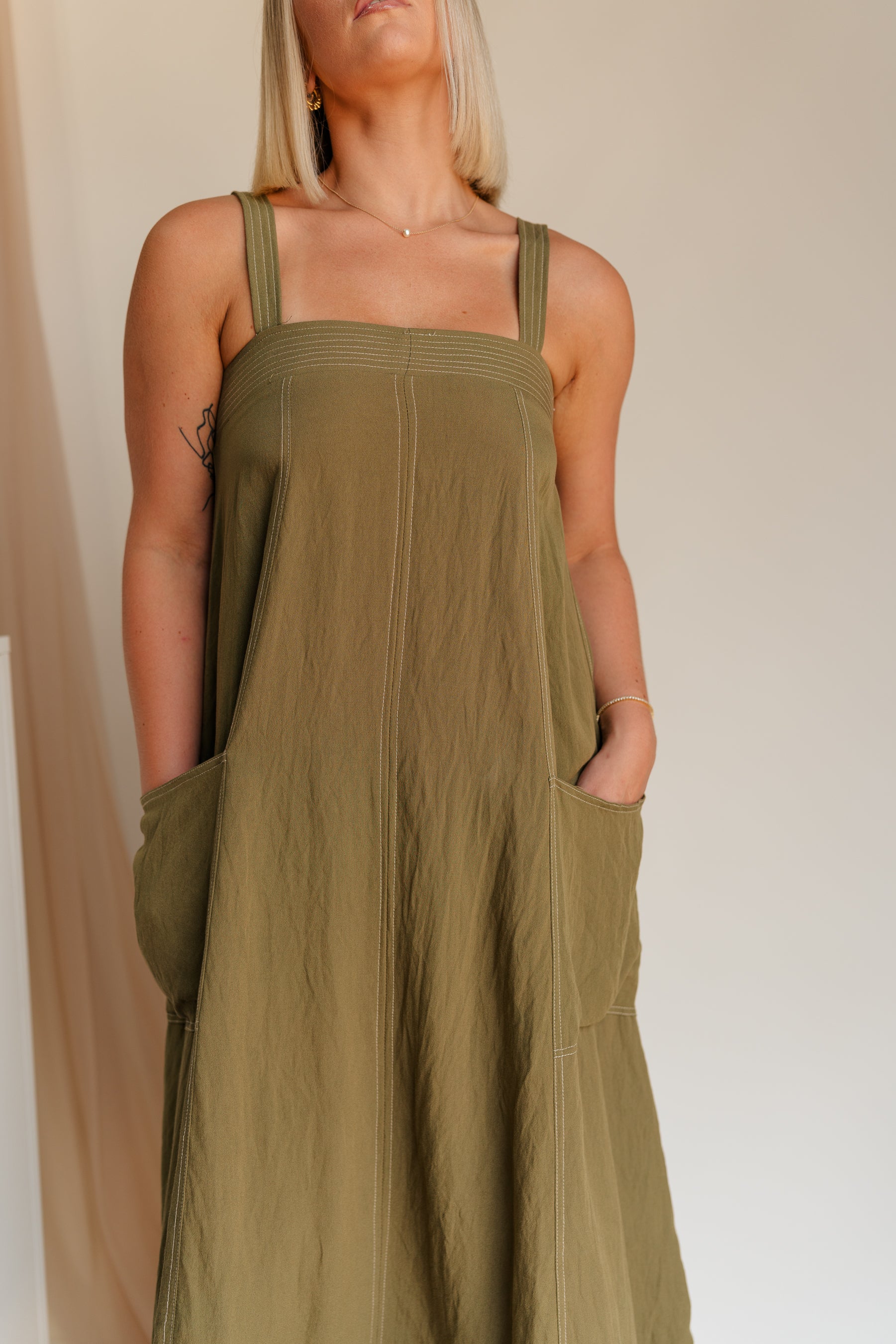 Close up of model wearing Melanie Olive Midi Dress. This is an olive midi dress, with white stitch details, midi length, pockets, and adjustable straps.
