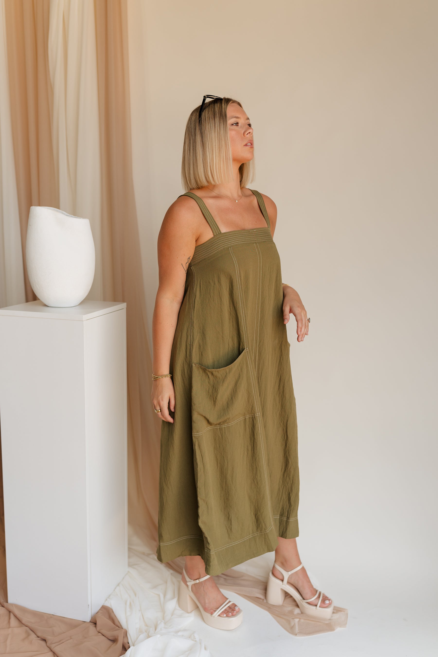 Side view of model wearing Melanie Olive Midi Dress. This is an olive midi dress, with white stitch details, midi length, pockets, and adjustable straps.