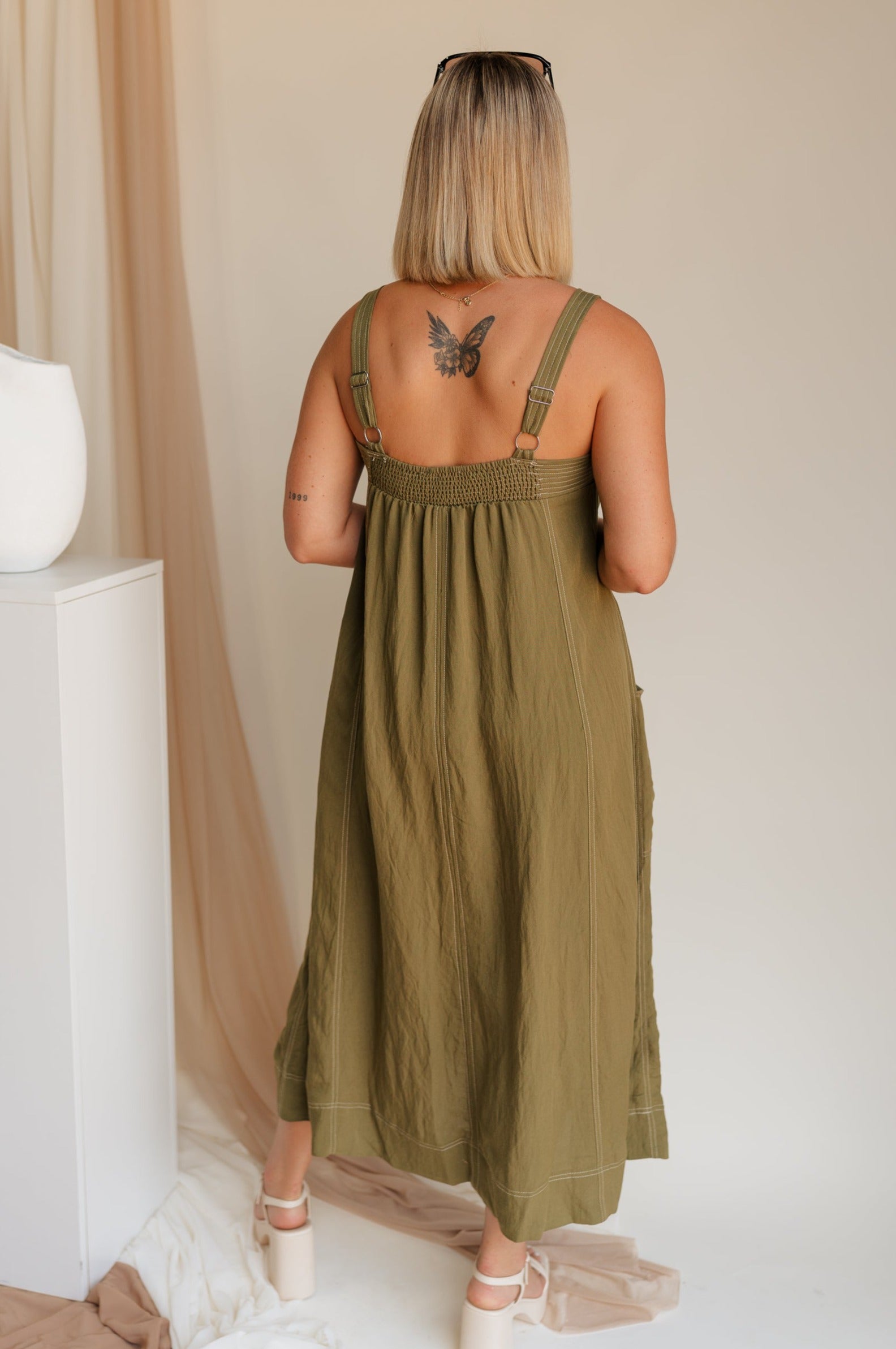 Back view of model wearing Melanie Olive Midi Dress. This is an olive midi dress, with white stitch details, midi length, pockets, and adjustable straps.