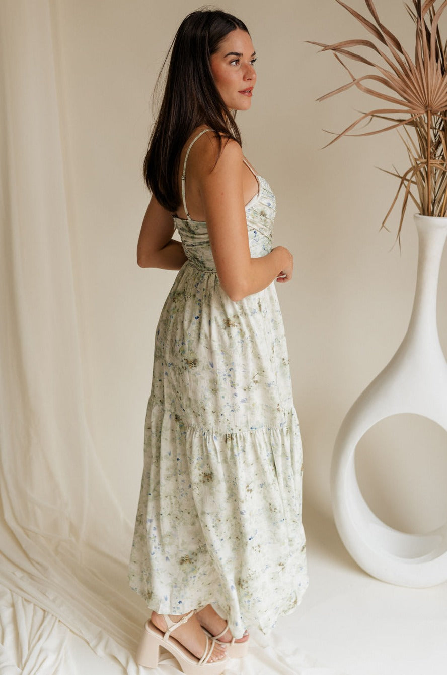 side view of female model wearing the Clara Sage & Blue Floral Midi Dress which features Green, Blue and White Floral Print, Midi Length, White Lining, Flare Hem Skirt, Ruched Criss Cross Upper Detail, Adjustable Straps and Monochrome Back Zipper with Hook Closure