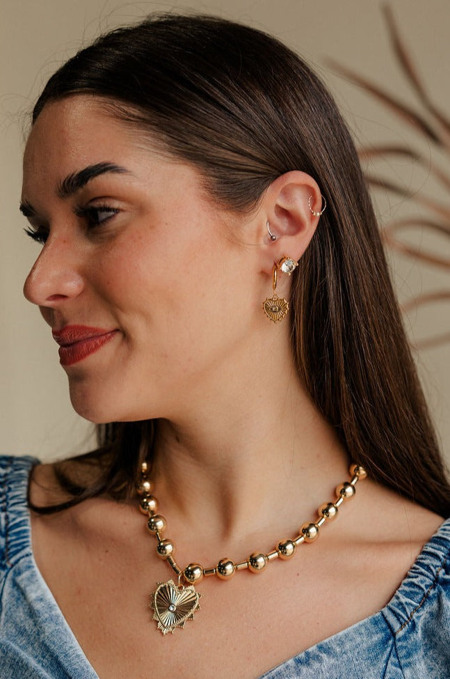 Side view of female model wearing the Vera Rhinestone & Gold Heart Shaped Huggie Earring which features heart shaped gold huggies with a clear stone detail