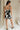full body back view of female model wearing the Lila Black & Cream Tie Straps Romper which features Black and White Geometric Pattern, White Shorts Lining, Two Side Pockets, Ruffle Upper, Square Neckline and Tie Straps