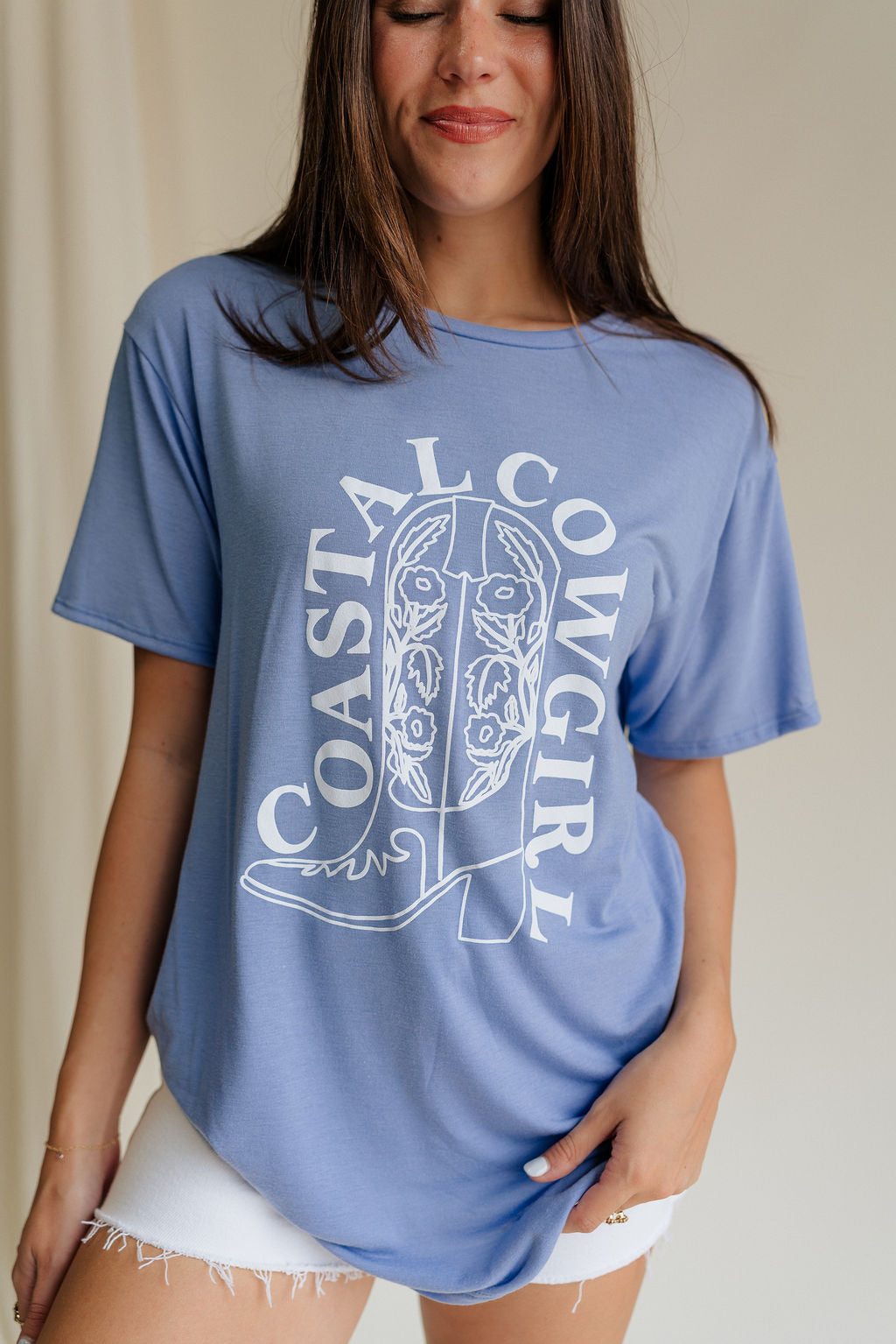 Upper body front view of female model wearing the Coastal Cowgirl Light Blue Graphic Tee that has light blue fabric, a white cowgirl boot graphic, white "Coastal Cowgirl" text, short sleeves, and a round neck.