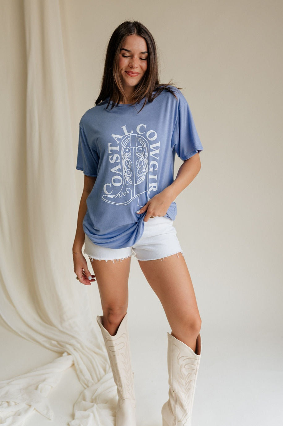 Full body front view of female model wearing the Coastal Cowgirl Light Blue Graphic Tee that has light blue fabric, a white cowgirl boot graphic, white "Coastal Cowgirl" text, short sleeves, and a round neck.