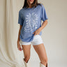 Full body front view of female model wearing the Coastal Cowgirl Light Blue Graphic Tee that has light blue fabric, a white cowgirl boot graphic, white "Coastal Cowgirl" text, short sleeves, and a round neck.